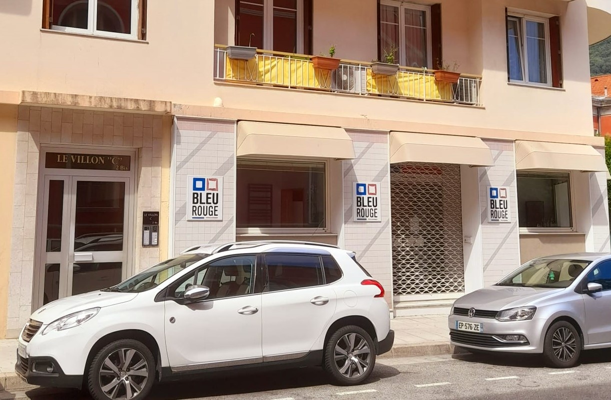 Façade d’immeuble beige avec balcons jaunes, volets rouges, auvents et voitures garées devant.