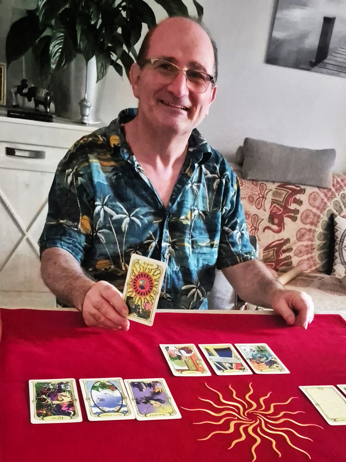 Homme souriant en chemise colorée tenant une carte, assis à un bureau avec livres et plante.